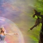 Princess in a silver ball gown and crown reaches toward a green-skinned witch on a broom, inside a large magical bubble against a colorful sky.